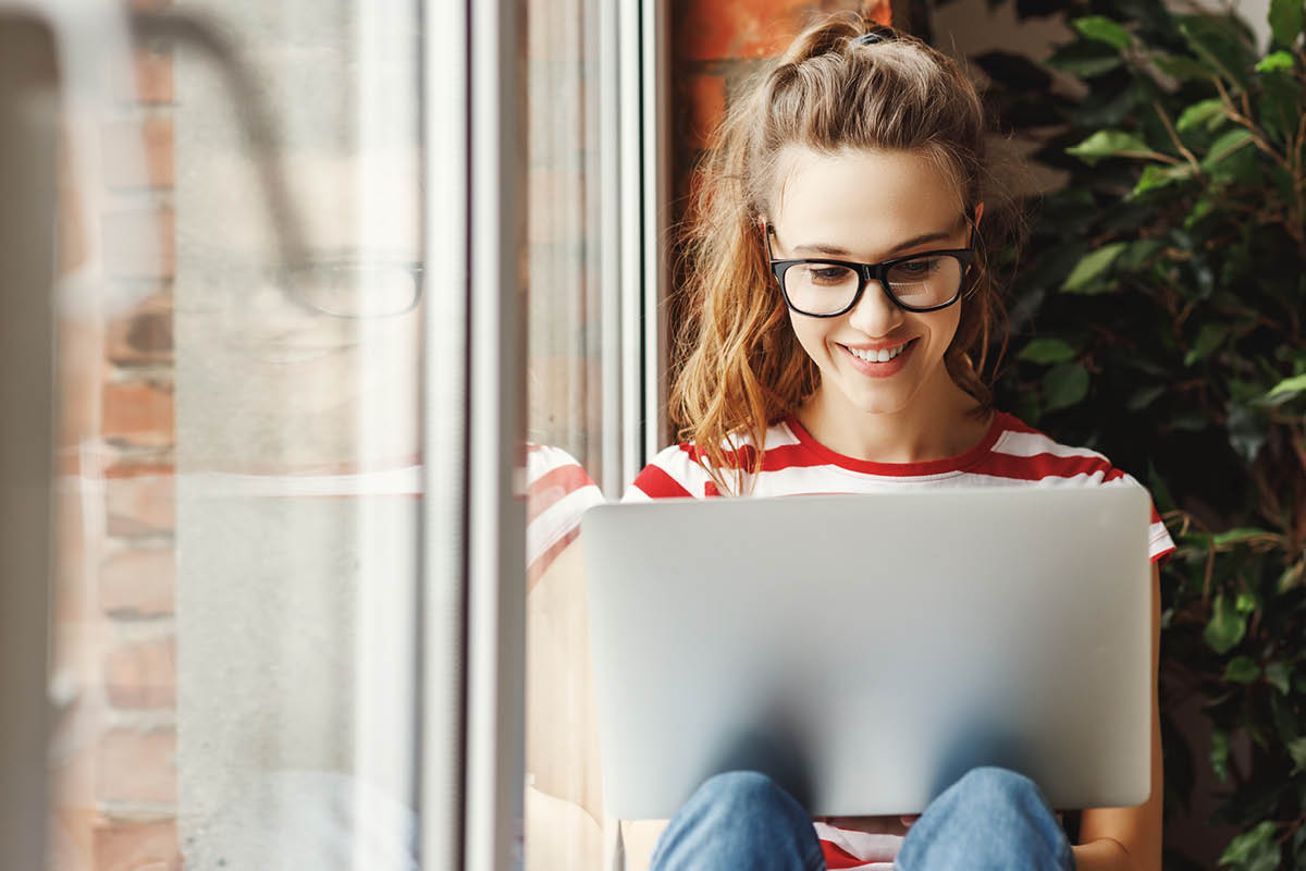 Junge Frau sitzt auf einer Fensterbank und recherchiert etwas mit ihrem Laptop